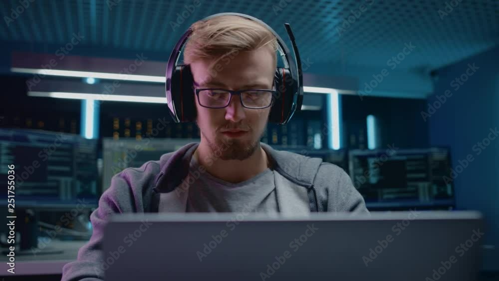 Aufkleber Portrait of Software Developer / Hacker / Gamer Wearing Glasses and Headset Sitting at His Desk and Working / Playing on Laptop. In Background Dark Neon High Tech Environment with Multiple Displays