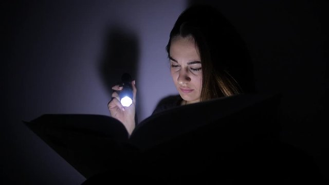 close-up portrait girl is reading a book with a flashlight in a dark room at night. 4k. Motion shooting