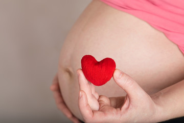Young pregnant woman keeps small red plush heart close to her belly.