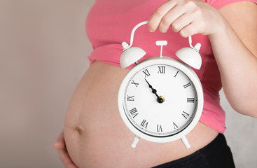 Young pregnant woman keeps vintage alarm clock close to her belly.