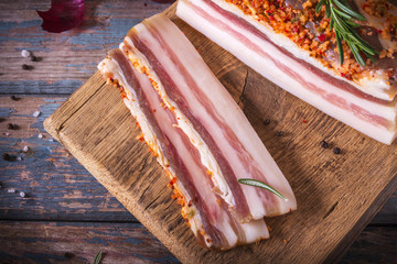 Chunks of salted bacon on dark background. Healthy food with spices, herbs is decorated rosemary on...