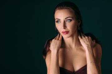 Portrait of magnificent sexy woman in evening dress with diamond earring posing over dark green background