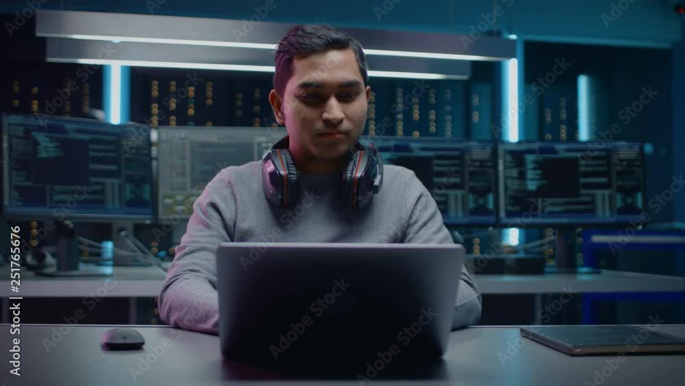 Poster Portrait of Software Developer / Hacker / Programmer Sitting at His Desk and Working on Laptop Computer In the Background Dark Neon High Tech Environment with Multiple Displays. 