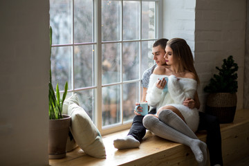 Fototapeta na wymiar beautiful girl and the guy are sitting on the windowsill near the window with a cup of tea and look out the window