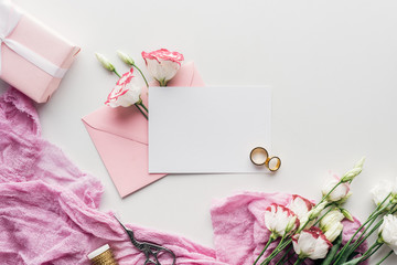 top view of empty card with pink envelope, flowers, cloth, wrapped gift, scissors and golden wedding rings on white background