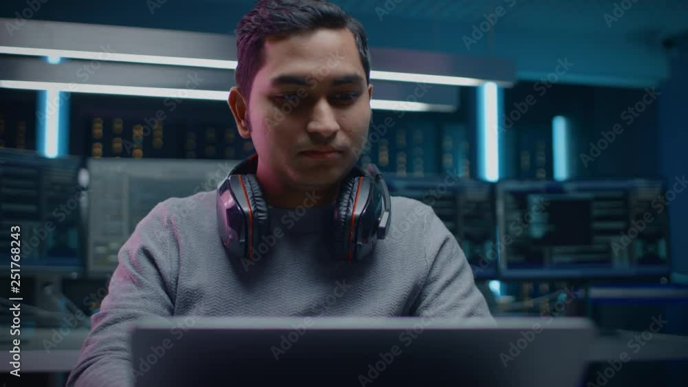 Aufkleber Portrait of Handsome Indian Software Developer / Hacker / Programmer Sitting at His Desk and Working on Laptop Computer In the Background Dark Neon High Tech Environment with Multiple Displays. 