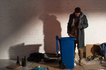homeless man standing near garbage container surrounded by rubbish
