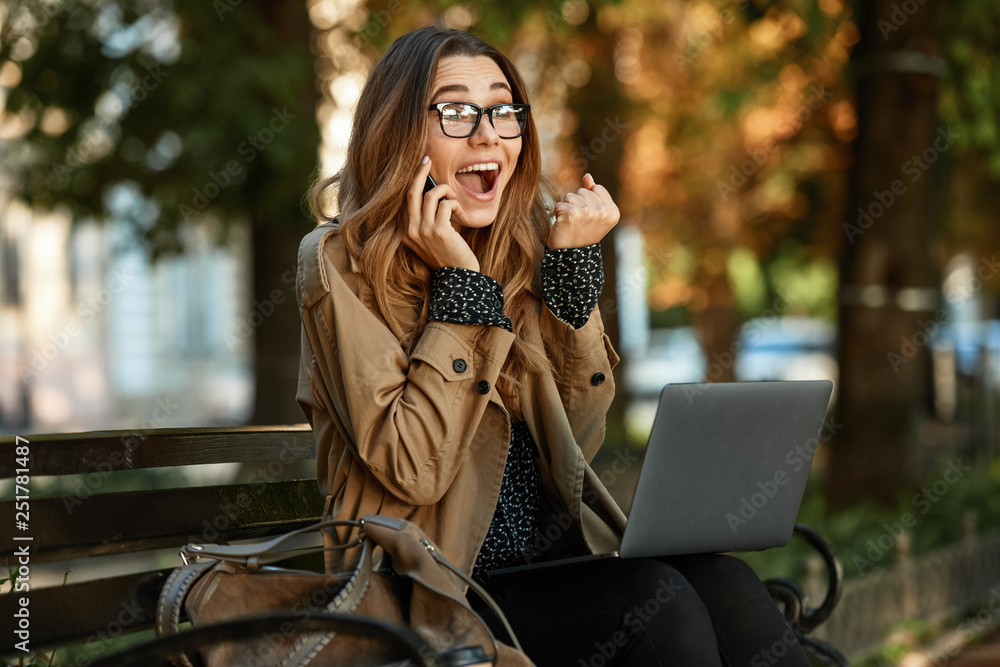 Sticker Photo of happy girl using mobile phone and laptop while sitting on bench in sunlit alley