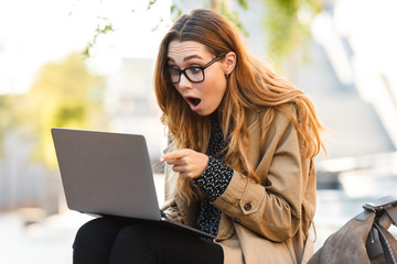 Photo of surprised woman working on laptop while sitting on bench in city street
