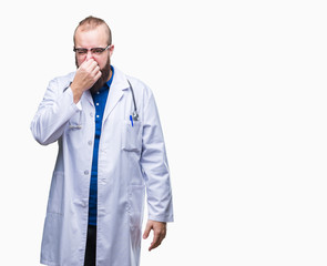 Young caucasian doctor man wearing medical white coat over isolated background smelling something...