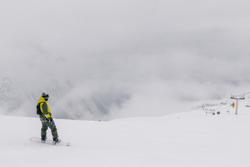 Snowboarder on top of the mountain before the start of the descent. Clouds lie below the ski lift station. Mountain gorge below in the fog