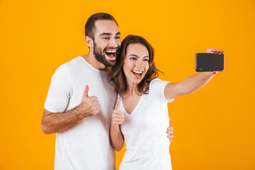 Portrait of two happy people man and woman taking selfie photo on smartphone, isolated over yellow...