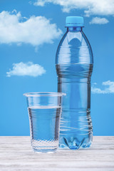 Bottle with water on white table and  blue background with clouds. Fresh water in plastic cup.Copy space. 