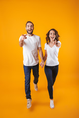 Full length photo of cheerful couple holding hands while walking, isolated over yellow background