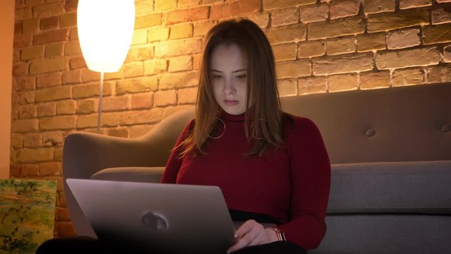 Closeup shoot of young attractive caucasian female using the laptop while sitting on the floor in a cozy apartment