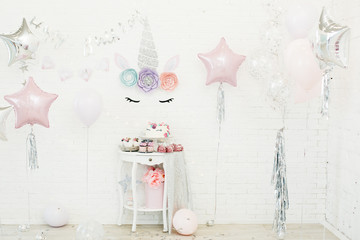 A table with sweets stands on the background of white brick walls: marshmallows, cupcakes, blueberry cake with chocolate cream, decorated under the head of a unicorn with a multi-colored mane, garland