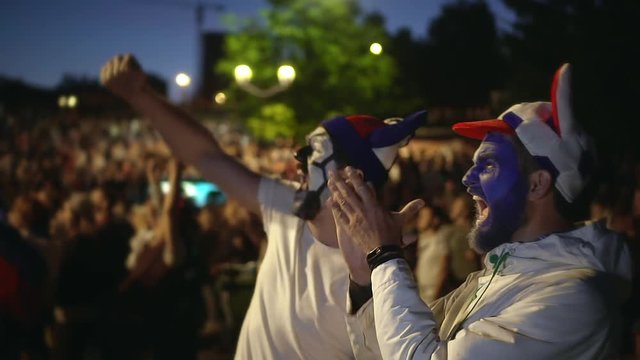 Football fan shouts, rejoices goal favorite team. Person paint face in delight victory match. Close up guy screaming furiously, jumping with friend against backdrop crowd winning football slowmotion.