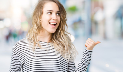 Beautiful young blonde woman wearing stripes sweater over isolated background smiling with happy face looking and pointing to the side with thumb up.