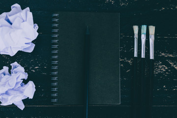 black notepad on dark desk with brushes and scrunched paper balls