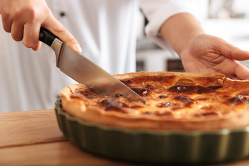 Image of european woman chef wearing white uniform cutting apple pie, while cooking in kitchen at the bakery