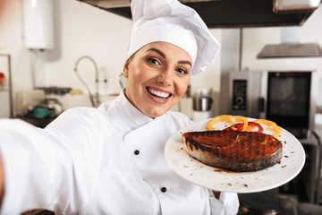 Portrait of positive woman chef wearing white uniform, taking selfie photo while holding plate with food in kitchen at the restaurant