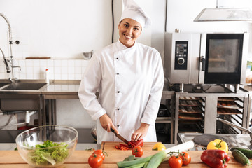 Photo of pretty woman chef wearing white uniform making salad with fresh vegetables, in kitchen at...