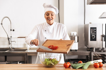 Photo of positive woman chef wearing white uniform making salad with fresh vegetables, in kitchen at the restaurant