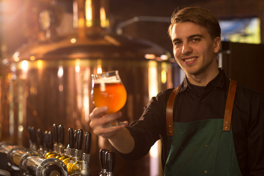 Charming bartender looking at camera, showing glass of delicious fresh beer and smiling. Handsome barman wearing in black shirt and apron. Brew bronzed kettle behind.
