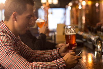 Side view of client of bar sitting at bar counter, holding beer glass in hand. Man smiling and looking at mobile phone. Man spending time in beer house or brewery, tasting delicious beer and resting.