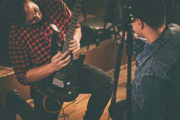 Guitarist playing electric guitar emotionally and professionally. Musician wearing in casual checked shirt, standing on knee. Singer in jeans jacket singing. Friends on band repetition.