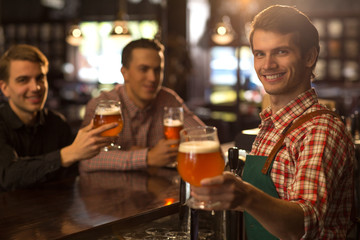 Positive bartender posing, smiling and showing beer. Handsome barman wearing in checked shirt and apron. Clients of beer house or brewery sitting with beer glasses in hands behind.