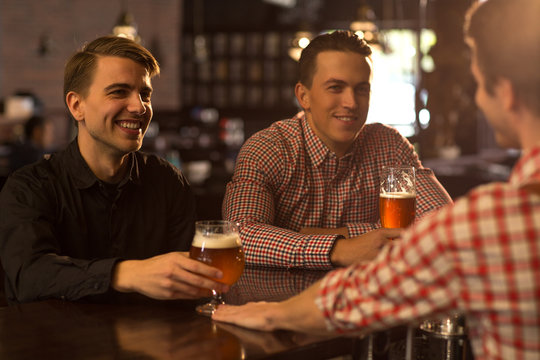 Bartender serving people in bar. Cheerful men sitting at bar counter, looking at barman, smiling and holding beer glasses. Friends hanging out together in beer house.