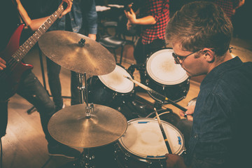 Fototapeta na wymiar Professional drummer practicing on repetition with other members of rock band in studio. Young male musician using drumsticks. Guitarists playing on instruments on background.