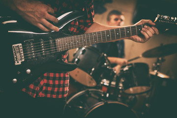 Close up of unrecognizable guitarist playing black modern electric guitar. Musician holding hand on guitar strings. Drummer drumming behind. Concept of music instruments, rock band repetition.