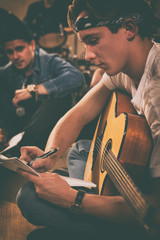 Side view of charming young musician sitting on floor with bass guitar. Guitarist in bandana writing with pen on papers, composing songs. Boys of rock band sitting behind.