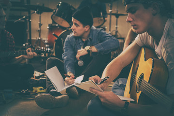 Handsome guitar with bass guitar holding papers and writing with pen. Members of rock band sitting on floor behind composing songs. Singer in black cap holding phone and looking away.