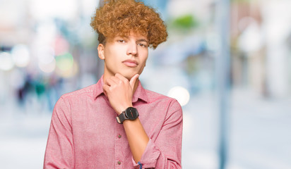 Young handsome business man with afro hair looking confident at the camera with smile with crossed arms and hand raised on chin. Thinking positive.