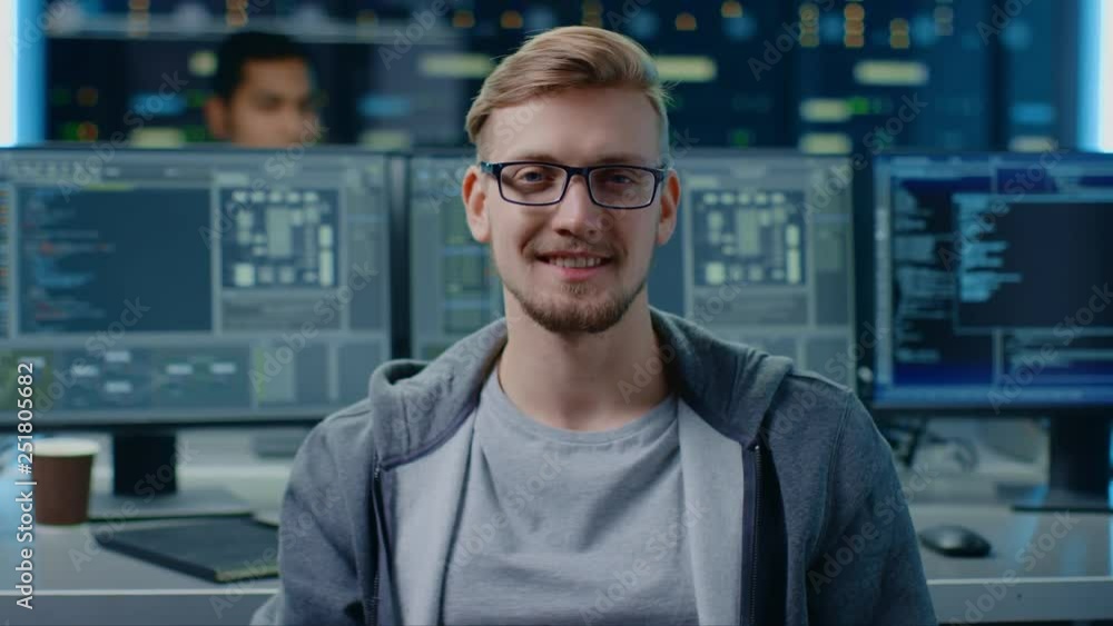 Poster Portrait of a Smart and Handsome IT Specialist Wearing Glasses Smiles, Behind Him Personal Computers with Screens Showing Software Program with Coding Language Interface in Data Center