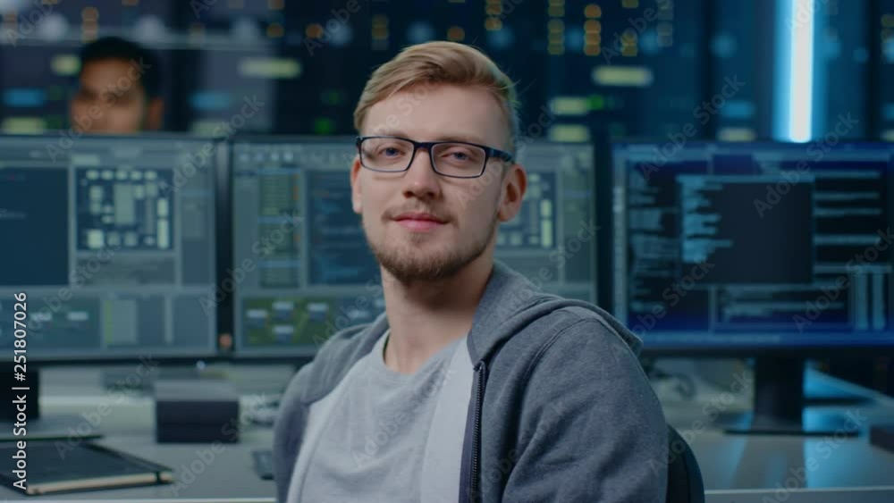 Poster Portrait of a Smart and Handsome IT Specialist Wearing Glasses Smiles at the Camera, Behind Him Personal Computers with Screens Showing Software Program