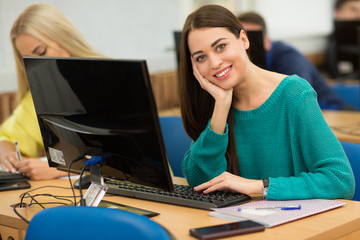 Smiling young girl, wearing in casual clothes, sitting at table with personal computer at...