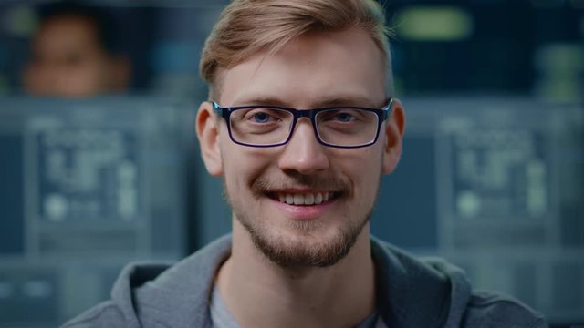 Portrait of a Smart and Handsome IT Specialist Wearing Glasses Smiles at the Camera. In the Background Unfocused Personal Computers with Screens in Technological Research Data Center
