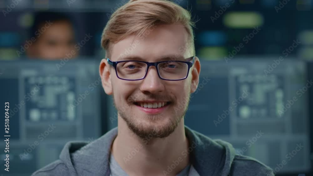 Aufkleber Portrait of a Smart and Handsome IT Specialist Wearing Glasses Charmingly Smiles at the Camera. In the Background Unfocused Personal Computers with Screens in Technological Research Data Center