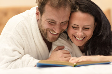 Closeup of laughing couple in white robes and reading funny story together. Cheerful woman and man lying on bed, hugging each other and looking at interesting book. Concept of entertaining at home.