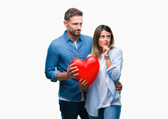 Young couple in love holding red heart over isolated background serious face thinking about question, very confused idea