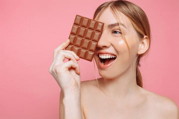 young woman with clean skin holding a black bar of chocolate in her hands, closing one eye with chocolate on a pink background