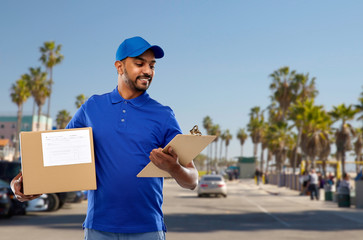 mail service and shipment concept - happy indian delivery man with parcel box and clipboard in blue uniform over venice beach background in california