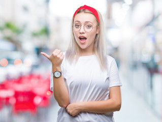 Young blonde woman wearing glasses over isolated background smiling with happy face looking and pointing to the side with thumb up.