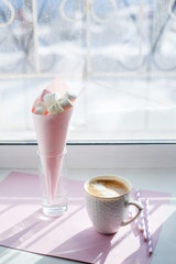 A white cup of coffee with pink marshmallow in the paper cone near the window. A warm drink for a cozy morning. breakfast or snack. metaphor for pausing at work or warm morning