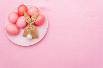 Easter eggs concept, table arrangement decoration. Pink (rosy) eggs with bunny (rabbit) paper gift egg wrapping on plate. Cloth (linen) background, top view