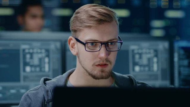 Portrait of a Smart and Handsome IT Specialist Wearing Glasses Works on a Personal Computer. In the Background Unfocused Personal Computers with Screens in Technological Research Data Center